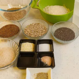 Divers bols contenant des farines, des graines, du psyllium, des épices et de l’eau, disposés sur un plan de travail clair pour préparer un pain aux graines sans gluten.